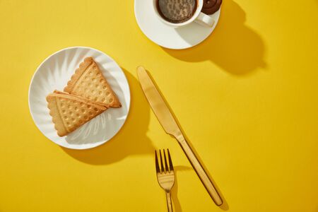 Top view of cup of coffee with golden cutlery and delicious cookies on yellow surfaceの写真素材