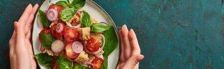 cropped view of woman holding plate with delicious Italian vegetable salad panzanella on textured green surface, panoramic shotの写真素材