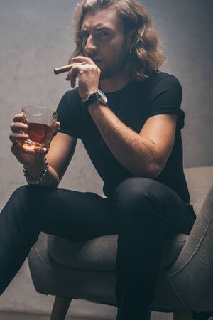 fashionable businessman in black outfit smoking cigar and drinking whiskey in armchair near grey wallの写真素材