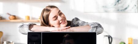 panoramic shot of smiling and attractive woman lying on microwave in kitchenの写真素材