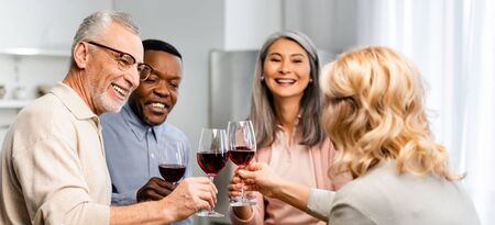 panoramic shot of smiling multicultural friends clinking with wine glasses in kitchenの写真素材