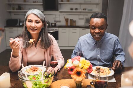 selective focus of smiling multicultural man and woman talking and eating pasta during dinner