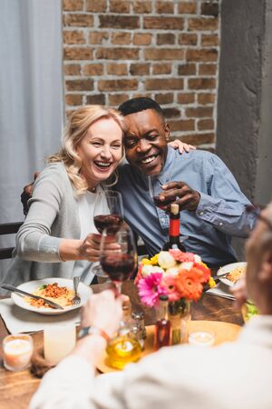 selective focus of smiling multicultural friends hugging and clinking during dinner