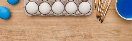 top view of blue watercolor paint in bowl near chicken eggs and paintbrushes on wooden table,...