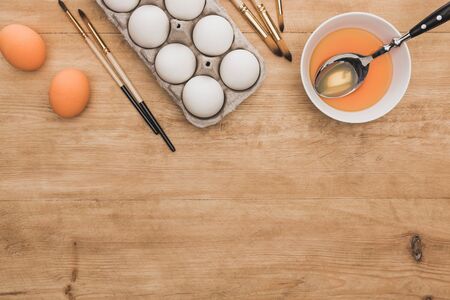 top view of orange watercolor paint in bowl near chicken eggs and paintbrushes on wooden table