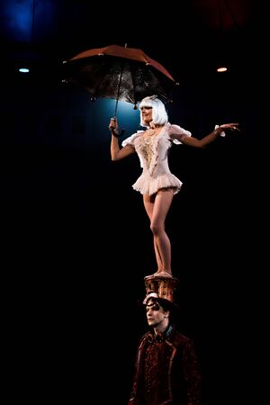 attractive woman with umbrella performing while standing on hat of man in circus