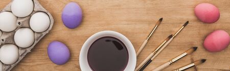 top view of watercolor purple paint in bowl near chicken eggs and paintbrushes on wooden table,...
