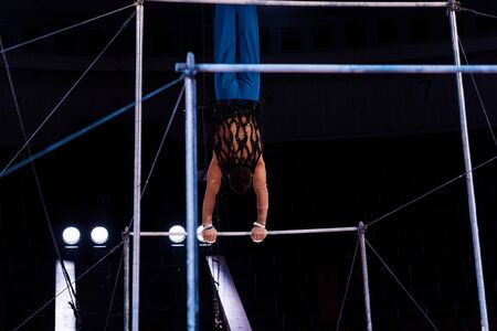 back view of athletic gymnast performing on horizontal bars in arena of circusの写真素材