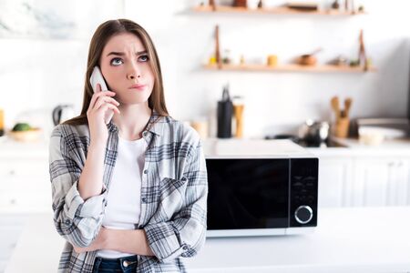 sad woman talking on smartphone near microwave in kitchenの写真素材
