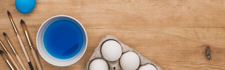 top view of watercolor blue paint in bowl near chicken eggs and paintbrushes on wooden table, panoramic shotの写真素材