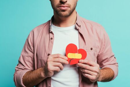 cropped view of upset man holding broken heart with plaster, isolated on blue