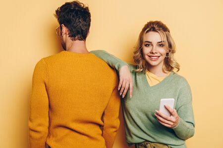 back view of man standing near smiling girl using smartphone on yellow background
