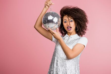 shocked african american girl in paillettes dress holding disco ball, isolated on pinkの写真素材
