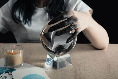 Cropped view of magician with black painted hand holding crystal ball by watercolor paintings and candle on table isolated on blackの写真素材