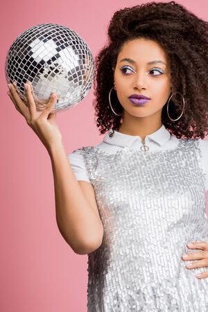 beautiful african american girl in paillettes dress holding disco ball, isolated on pink
