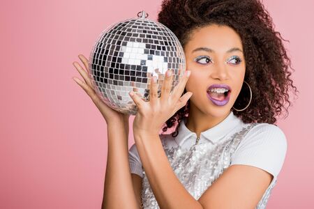 excited african american girl in paillettes dress holding disco ball, isolated on pink