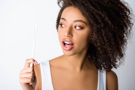 surprised african american woman with dental braces looking at toothbrush, isolated on grey