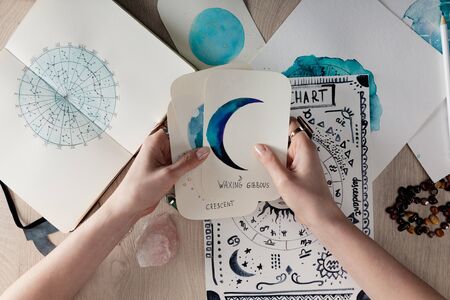 Top view of astrologer holding watercolor paintings with moon phases on cards by birth chart on...
