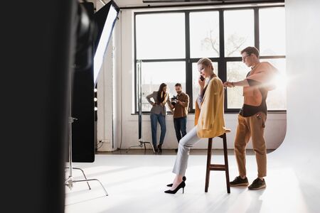 stylist using lint roller, model talking on smartphone, producer and photographer talking on...