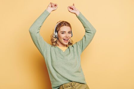 cheerful girl listening music in wireless headphones, dancing and looking at camera on yellow...