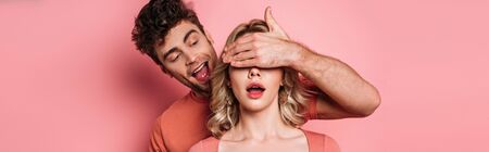 panoramic shot of tricky man covering eyes of surprised girl with hand on pink background