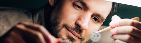 Panoramic shot of handsome watchmaker looking at watch part by lampの写真素材