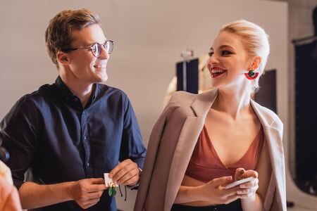 smiling stylist holding earrings and looking at stylish model with smartphone on backstage