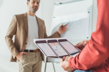 Selective focus of designer holding ux mobile application layout with colleagues beside whiteboard...