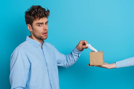 cropped view of woman giving pack of paper napkins to upset man looking at camera on blue background