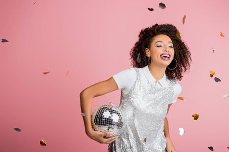 smiling african american girl in paillettes dress holding disco ball, isolated on pink with confetti