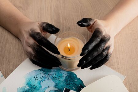 Cropped view of witch with black paint on hands holding candle by cards with watercolor zodiac signs...