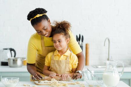 cute african american kid holding rolling pin near cheerful mother and raw dumplings on cutting boardの写真素材