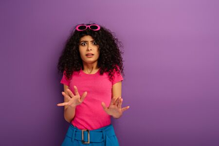 scared bi-racial girl showing stop gesture while looking at camera on purple background
