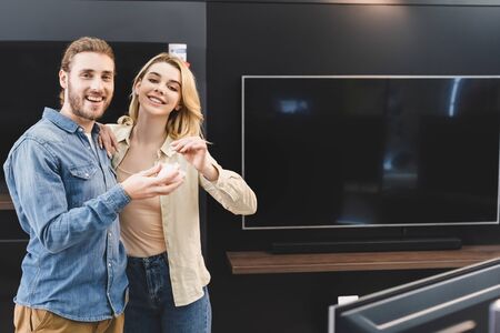 smiling boyfriend holding piggy bank and girlfriend putting coin into it in home appliance storeの写真素材