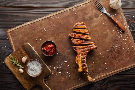Top view of delicious ribeye steak with tomato sauce, garlic and salt on stone board on wooden...
