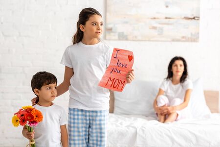 cute brother and sister holding i love you mom card and flowers, while mother sitting in bed