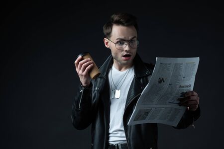 shocked stylish brutal man in biker jacket with newspaper and coffee to go isolated on black
