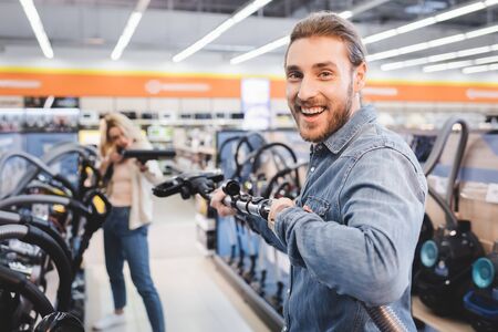 selective focus of smiling boyfriend holding vacuum cleaner and girlfriend on background in home...