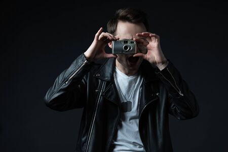 stylish brutal man in biker jacket taking picture on film camera with open mouth isolated on black