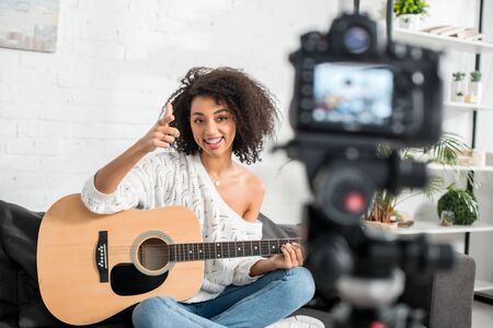 selective focus of cheerful african american girl in braces holding acoustic guitar and pointing with finger at digital cameraの写真素材