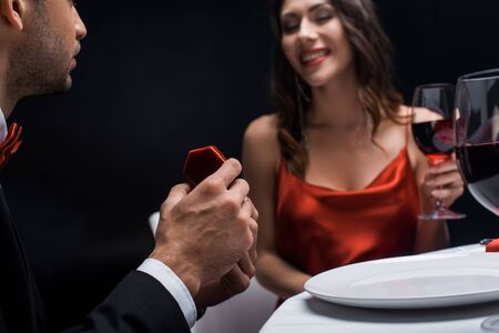 Selective focus of man presenting jewelry box at smiling girlfriend with wine glass during romantic dinner isolated on blackの写真素材