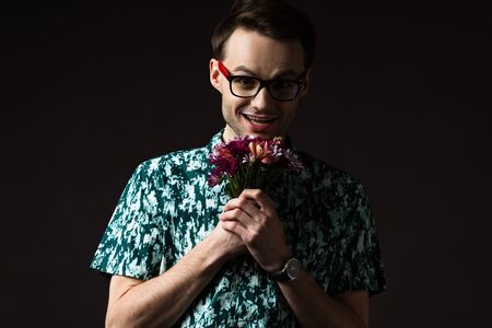 excited fashionable man in eyeglasses in blue colorful shirt holding floral bouquet isolated on blackの写真素材