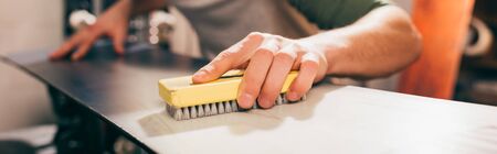 panoramic shot of worker using brush on snowboard in repair shop