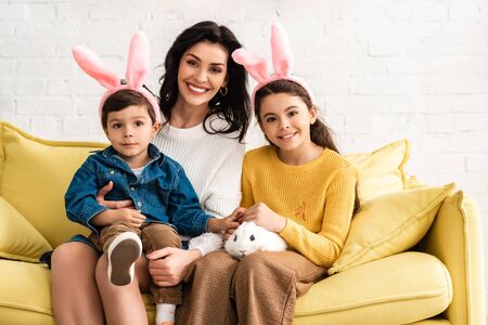 happy woman and cheerful children in bunny ears smiling at camera while sitting on yellow sofa with...