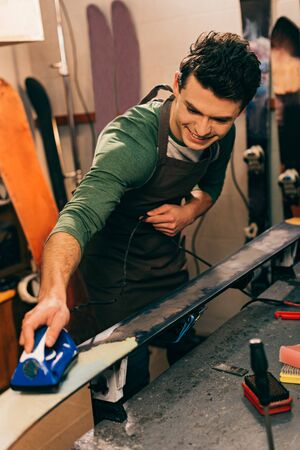smiling worker waxing ski with wax iron in repair shopの写真素材