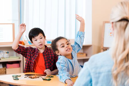 Selective focus of children with raised hands and teacher during lesson in montessori classの写真素材