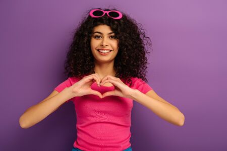 happy bi-racial girl showing heart sign with hands while looking at camera on purple background