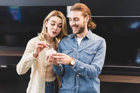 smiling boyfriend holding piggy bank and girlfriend putting coin into it in home appliance store