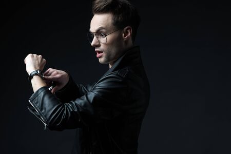 side view of shocked stylish brutal man in biker jacket pointing with finger at wristwatch isolated...