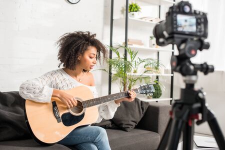 selective focus of curly african american girl playing acoustic guitar near digital cameraの写真素材